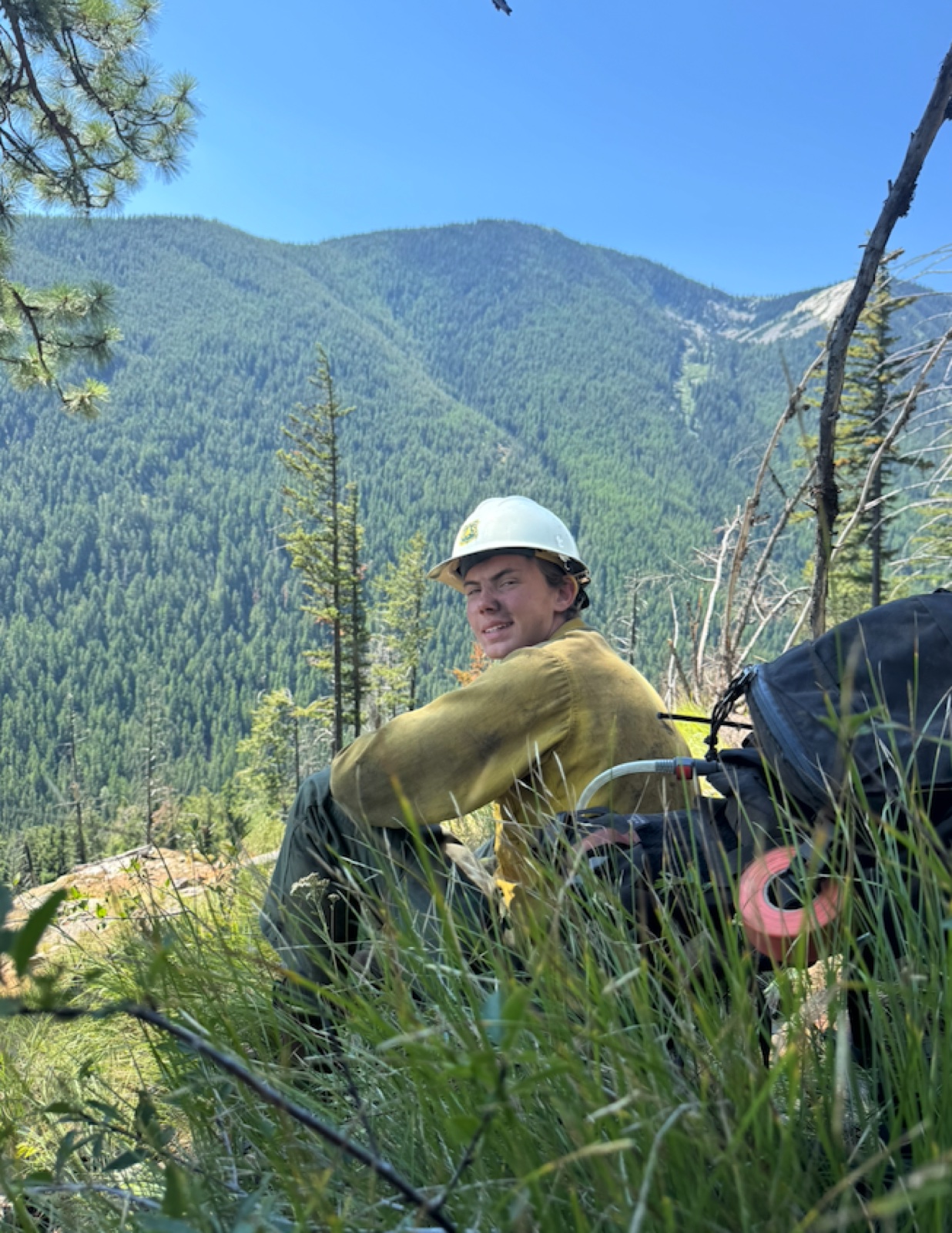 A wildland firefighter in a white helmet and yellow shirt sits on a grassy slope above a forested mountain valley